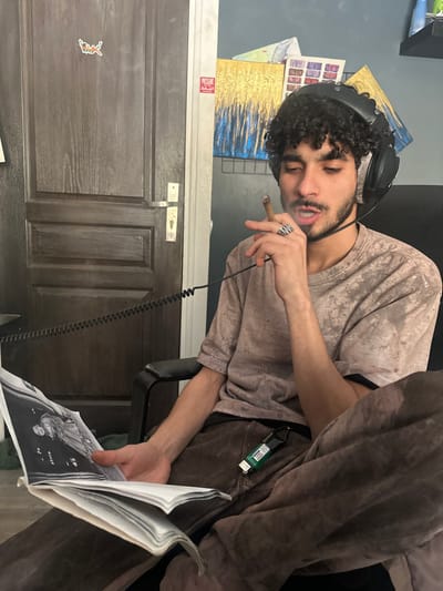 The scene is an indoor environment located in Marcoussis, France. A young adult male, characterized by dark curly hair and a beard, is seated in a dark-framed chair. He wears circumaural headphones connected by a coiled cable, a light brown mottled t-shirt, and dark brown trousers. A soft, brown fabric covers his lower extremities. A silver-toned ring is worn on the ring finger of his left hand. He holds a lit cigar or blunt near his mouth, from which smoke is visibly emanating, and his mouth is slightly open. In his right hand, resting on his lap, he holds an open book displaying a black and white photograph of two individuals. A green and white keychain or device is clipped to his right trouser leg. The immediate background on the left features a dark wooden two-panel door equipped with a metallic handle and a small, stylized winged figure sticker. To the right, a blue-gray wall is adorned with two framed abstract artworks exhibiting gold and blue textures, several smaller pinned papers, and a red and white sticker partially displaying the word "PRESSURE." An upper shelf with unidentifiable objects is visible in the top right corner.