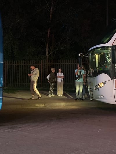A group of people stand outside a white bus near a fence at night in Terespol, Poland. One person is on their phone. Another person is shouting. The scene is lit by artificial lights, and the background is dark with trees.