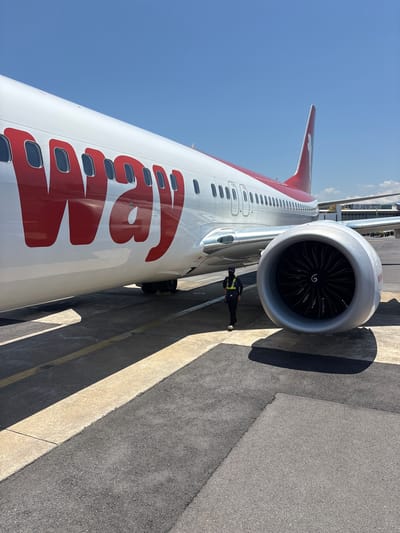 This is a photograph of an airplane parked on an airport tarmac under a clear blue sky. The perspective is from a low angle, looking up at the side of the aircraft and its engine. The airplane is predominantly white with large red lettering that appears to spell "JEJU" along its fuselage. The tail of the plane is also red. The engine, a prominent feature in the center of the frame, is large and silver with black fan blades visible inside. A person wearing a high-visibility vest and dark clothing is standing near the engine, appearing to be working or inspecting the aircraft. The tarmac is gray asphalt with yellow and white markings, and the bright sunlight casts sharp shadows, indicating it is daytime with fair weather. In the background, some airport structures and possibly other aircraft are visible. The overall scene suggests a normal operational day at an airport.