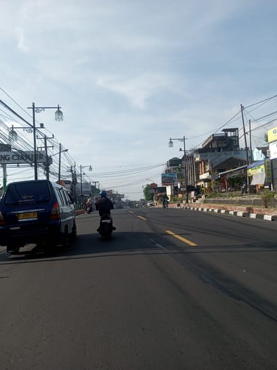 The image shows a street scene in an urban environment. A dark blue minivan is on the left, displaying a license plate with the number "F 1901 ZF". A motorcyclist is positioned ahead of the minivan, also traveling in the same direction. The road has a yellow line marking its center. On the right side of the road, there are buildings, various signs, and what appears to be a curb painted with alternating black and white sections. Power lines are visible overhead, crisscrossing the sky. Street lamps with decorative designs are also present. The sky is light blue with scattered clouds. It is not in Phou Khoun, Laos. A sign in the image says "Cianjur".