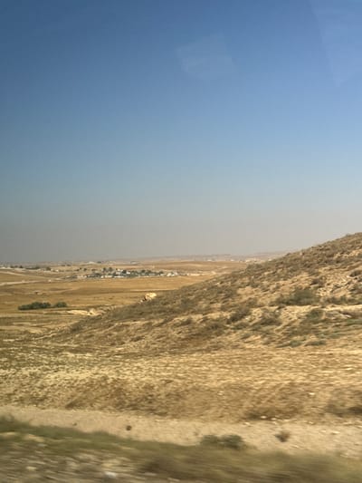 This media file depicts an aerial or elevated view of a dusty landscape under a clear blue sky, transitioning to a hazy horizon. The scene is dominated by a rugged, brown mountainside on the right, with sparse vegetation clinging to its slopes. The left side of the image is a vast expanse of pale blue sky, hinting at a bright day.
In the distance, below the hazy horizon, a small settlement or village is visible, characterized by a cluster of buildings. The terrain surrounding the settlement appears arid and flat, with some signs of dry fields or undeveloped land. There are no visible people or specific objects that stand out, making the scene feel expansive and somewhat desolate.
The time of day appears to be daytime, likely late morning or early afternoon, given the brightness of the sky. The presence of haze suggests dry or dusty atmospheric conditions, possibly due to wind or arid climate. The location cues, such as the arid landscape and the style of settlement, align with regions like al-Qasoum Regional Council in Israel. The overall impression is one of a remote, natural environment with a small human presence. No text is visible in the image.