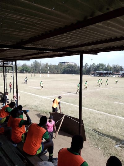 The image depicts a daytime view of a sports field within the city of Nakuru, Kenya, observed from beneath a covered spectator stand.

In the foreground, a corrugated metal roof, supported by dark metal beams and posts, extends over a seating area with wooden benches. Multiple individuals, predominantly male, are seated or standing under the cover. Several are wearing orange vests over green shirts and dark shorts; one male in an orange vest is seated and drinking from a blue plastic bottle. A male in a black cap and orange vest stands near a dark barrier on the right side of the frame. Below the main seating area, a female in a pink shirt and black cap holds a long, flat wooden object while standing next to a makeshift wooden barrier.

The midground features a large, open grass field with white line markings, indicative of a sports pitch. The grass appears somewhat worn. Two sets of white H-shaped rugby goalposts are prominently visible at opposing ends of the field. Scattered across the field are numerous individuals, approximately 15-20, dressed in light green or yellow jerseys with darker green shorts and socks. They are in various states of movement, suggesting a rugby match is in progress or paused.

The background shows a dense line of green trees bordering the far side of the field. Above the tree line, several buildings, including a multi-story structure, are visible in the distance. A tall light standard is also present on the right side of the background. Further right, a collection of people and vehicles suggests additional spectator areas or a parking zone. The sky above is largely clear with some diffused light, indicating daytime conditions.