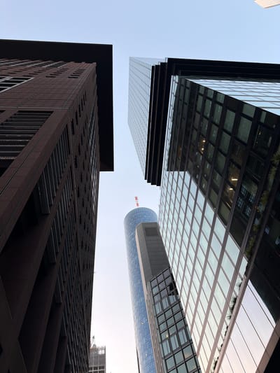The image, captured from a low-angle perspective, showcases the towering skyscrapers of Frankfurt, Germany's financial district. The foreground features a dark, rectangular building on the left, its facade constructed with dark reddish-brown brickwork and a repeating pattern of dark rectangular openings. To its right, a modern skyscraper with a glass facade dominates the view, its reflective surfaces mirroring the sky and other buildings. In the mid-ground, a cylindrical building with a blue glass exterior is visible, topped with a red and white antenna. Further in the background, another glass skyscraper and a construction site with a crane are discernible. The sky is clear and pale, suggesting either early morning or late afternoon. There are no people visible in the scene.