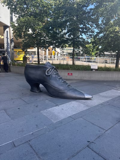 An outdoor, ground-level, medium shot captures a large, dark gray metal sculpture of a high-heeled shoe situated on a gray paved surface in Burnaby, Canada. The shoe, detailed with laces, eyelets, and stitching, is positioned at a slight angle, with its toe pointing towards the right. In the background, lush green trees with dappled sunlight filtering through their leaves frame the scene. Behind the trees, a concrete wall runs horizontally, with a sign that reads "NO LOITERING" in red lettering mounted on it. Further in the background, a yellow bus with the words "Desserts d spot" is visible, alongside other urban elements like railings, streetlights, and buildings. The pavement is made of large, rectangular gray tiles, some of which appear slightly wet, suggesting recent rain or cleaning.