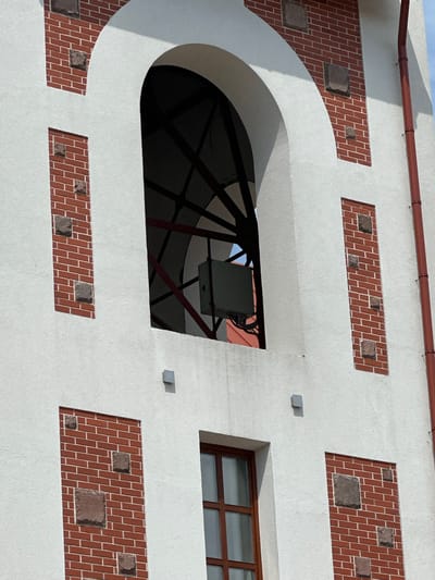 The image displays a detailed section of a building facade, characterized by white textured plaster or stucco surfaces contrasted with decorative red brick rectangular panels. These brick panels are intermittently adorned with roughly square, darker stone inserts.
Prominently featured in the upper central portion is a large, deeply recessed arched opening, which is dark within and reveals a structural metal framework. Mounted inside this framework is a light-gray rectangular equipment enclosure, from which dark cabling is visibly trailing. Through the arch, a sliver of light-blue sky and a faint reddish-orange surface are discernible in the background.
Below the arched opening, a smaller rectangular window with a brown, multi-pane wooden frame is present. To the right side of the facade, a reddish-brown vertical downspout runs from top to bottom, secured by lighter-colored brackets. Several small, light-gray square fixtures are sparsely mounted on the white plaster sections of the wall. The scene is illuminated by bright daylight, casting subtle shadows.
This architectural detail is observed on a building located in Бараўлянскі сельскі Савет, Belarus.