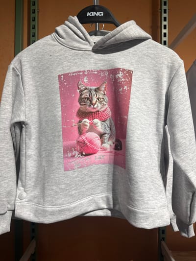 A light grey melange hooded sweatshirt hangs on a black plastic hanger marked with "KING" in white uppercase letters. The garment, featuring long sleeves and a cuffed hem, is displayed in a retail environment within the city of Perugia, Italy. A small, circular, light-colored button is visible on the lower left side of the sweatshirt. The front of the hoodie presents a rectangular graphic print with a distressed pink background. This graphic depicts a domestic tabby cat, characterized by grey and brown striped fur, seated upright. The cat wears a thick, pink knitted scarf around its neck and holds a slender, light-colored object, consistent with a knitting needle, in its front paws, positioned above a loop of pink yarn. A spherical ball of matching pink yarn rests beside the cat to its lower right. The word "LAY" is faintly visible in white text near the bottom center of the graphic. Out-of-focus metal shelving units, appearing in shades of green and brown, are visible in the background, confirming an indoor commercial setting.