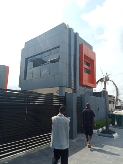 The image shows a modern building with a gray facade and an orange accent wall, located in Ujevwu, Nigeria (latitude: 5.472, longitude: 5.817). Two men are walking away from the building, one in a light-colored denim vest and black pants, the other in a black t-shirt and shorts. The man in the vest is looking at his phone. In the foreground, there is a black metal gate with horizontal bars. The building has large glass windows and a modern design. The ground is paved with gray concrete pavers. There is a small palm tree in the background. The sky is clear with a few clouds. The image is taken from a low angle, providing a wide perspective of the building.