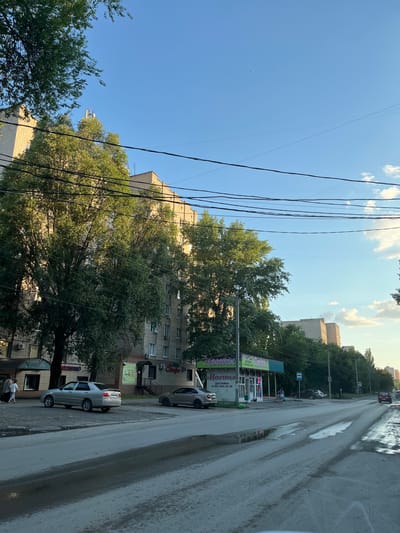 The image depicts a street scene in Taganrog City District, Russia. The photo is taken from a low angle, looking up at a multi-story building and a large tree with dense foliage. Overhead, a network of black power lines crisscrosses the frame against a clear blue sky dotted with white clouds.
In the foreground, a paved road runs along the right side of the image, appearing wet with visible puddles, suggesting recent rain. Several cars are parked along the street, including a silver sedan and a darker vehicle. A small storefront with a green awning and illuminated signage is visible. The text on the awning reads "Цветы" (Flowers) in large, bold letters, with additional smaller text below indicating "Доставка 8-924-822-12-29" (Delivery 8-924-822-12-29).
Further down the street, more buildings and trees are visible, creating a typical urban residential environment. Two people are standing near the storefront, their figures small in the frame. The lighting suggests it is daytime, likely late afternoon, given the warm tones and shadows. The overall atmosphere is calm and ordinary, capturing a moment of everyday life on a street in a Russian city.