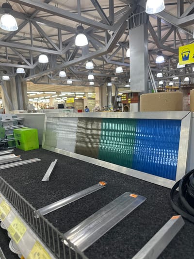 The image depicts the interior of a retail store, likely a hardware or building supply store, with high ceilings and industrial lighting. In the foreground, a dark gray, carpet-like surface displays various building materials. Prominently featured is a framed panel of ribbed translucent material, transitioning in color from white to green to blue. To the left of this panel is a small, bright green box with the "SUNLITE" logo. Several pieces of flat, metallic material, possibly aluminum or steel profiles, are arranged on the surface. In the background, shelves stocked with various goods are visible, along with a person in the distance. The overall environment suggests a commercial setting with an emphasis on construction or renovation supplies. The location is specified as the city of Dub, Montenegro.