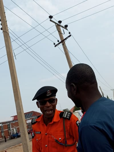The image depicts an outdoor scene with two men in conversation, with power lines and utility poles dominating the background. One man, in the foreground and on the left, wears an orange uniform with a black beret and sunglasses, suggesting he is a law enforcement officer. His uniform bears patches that read "NPF" and "NIGERIA POLICE". He has his mouth open as if speaking. The other man, on the right, is seen from behind, wearing a blue shirt. They appear to be standing on a dirt or sandy surface. In the background, a red brick building with glass windows and a sloped roof is visible, possibly a commercial establishment or government building. A blue tricycle and other vehicles are parked near the building. The sky is overcast with a light gray hue, and the overall setting suggests a slightly dusty environment. The location is identified as Tumfure, Nigeria.