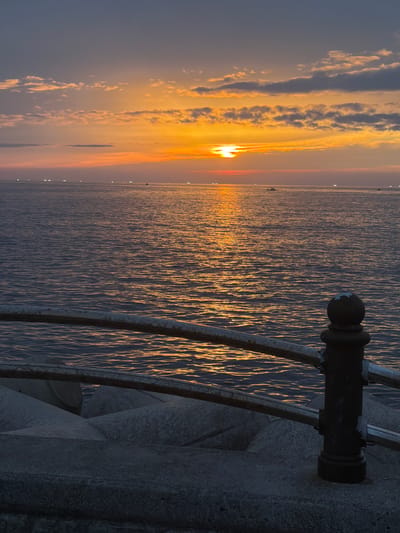 The image shows a sunset over the ocean with a view of the coast. The sky is a gradient of blues, oranges, and pinks, with clouds illuminated by the setting sun. The ocean surface is calm, reflecting the colors of the sky. In the foreground, there is a concrete barrier and a metal railing, suggesting a man-made structure along the coast. Several small boats are visible in the distance on the water, and lights are also visible along the horizon, hinting at a coastal town or city. The overall scene depicts a serene coastal sunset.