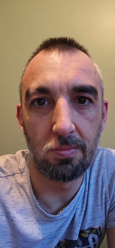 A close-up, rotated portrait shows a man with a full beard and short hair, looking directly at the camera. The image is oriented with the man's head tilted significantly, making it appear as if he is lying on his side or the photo was taken from a low, upward angle and then rotated clockwise. He has dark eyes and thick, dark eyebrows. His beard is dark with a noticeable amount of grey and white hairs, particularly around his chin and mustache. His hair, dark and short, shows signs of a receding hairline, and some grey is visible around the temples. His lips are full and have a reddish-purple hue. The man's skin tone is fair, with some minor blemishes or scars visible on his forehead/eyebrow area.
He is wearing a light blue, heathered t-shirt. A dark, patterned design, possibly floral or abstract, is visible on the lower left side of his shirt. A thin, metallic chain can be seen peeking out from under the collar of his shirt on the left side of his neck. The background is a plain, light greenish-yellow wall, suggesting an indoor setting. The lighting appears to be artificial, casting soft highlights and shadows on his face. The man's expression is neutral to slightly serious. No text is visible in the image. The photo was taken in Norwich, United Kingdom.