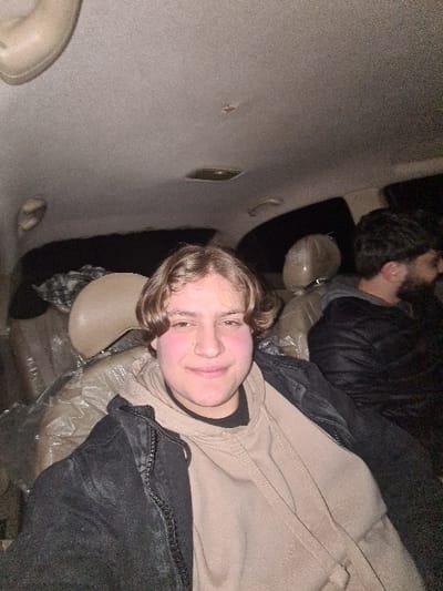 Two people are lying in the back of what appears to be a car. The person closest to the camera is light-skinned with light brown hair and a tan hoodie. He is looking at the camera and smiling. The other person has dark hair and is wearing a black jacket. They both appear to be on top of a makeshift bed made of blankets. The car interior is old and worn.