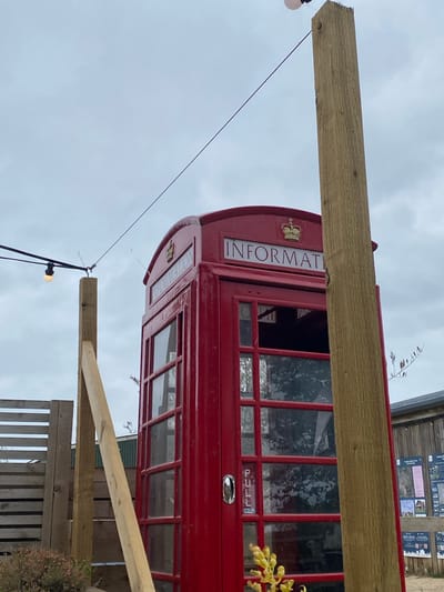 The image shows a red British telephone box, labeled "INFORMATION" with a crown emblem, partially obstructed by wooden beams, presumably part of a structure. The background includes a cloudy sky and a wooden structure with information boards. There is a string of lights with illuminated bulbs near the telephone box. The location is likely South Oxfordshire, United Kingdom, given the presence of the British telephone box and the style of signage.