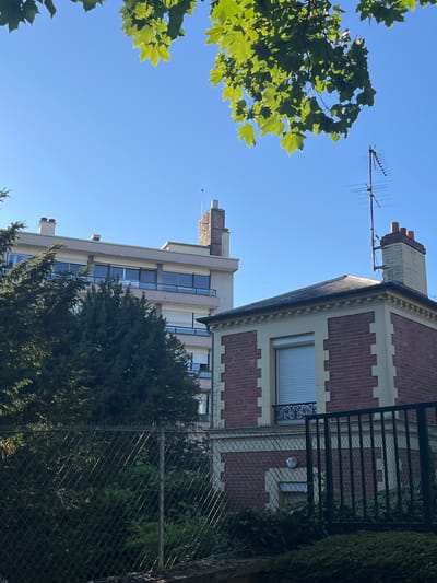 The image shows a clear blue sky above a residential area in L'Haÿ-les-Roses, France. In the foreground, a wire mesh fence partially obstructs the view of lush green trees and bushes. Beyond the fence, a two-story brick house with cream-colored trim and a dark roof is visible. The house has a prominent chimney with three terracotta pots and an antenna attached to the roof. To the left of the brick house, a taller, multi-story building with a light-colored facade and horizontal windows is seen, also featuring chimneys and an antenna. Green foliage from maple trees drapes into the frame from the top, illuminated by sunlight. The scene suggests a sunny day in a suburban or urban environment.