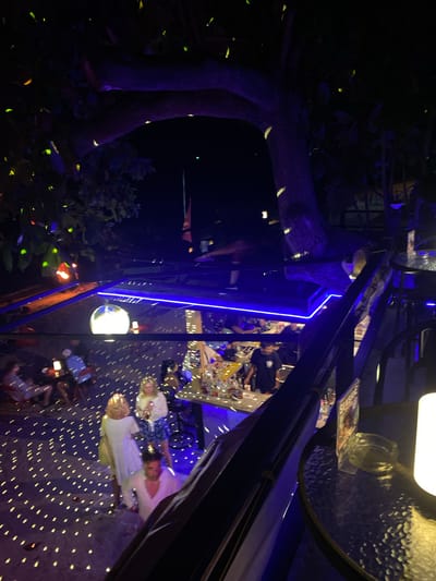 The image depicts an outdoor bar or restaurant setting at night in Ban Haad Rin, Thailand. Tables are visible, some with customers seated and interacting. There are people wearing light-colored clothing. The environment is decorated with lighting, including blue neon lights, overhead lamps, and a disco ball that projects light patterns on the ground. A tree and foliage are present outside the bar area, with some illuminated areas suggesting outdoor lighting.