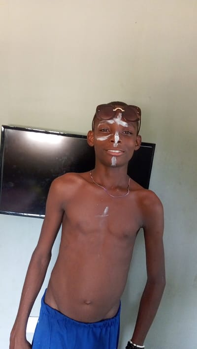 A shirtless, slender, dark-skinned young man stands in front of a wall with a television. He wears sunglasses on his head and blue shorts. White paint marks adorn his forehead, nose, and chest. He also wears a silver chain necklace and bracelets on his left wrist. The background consists of a plain wall, suggesting an indoor setting.