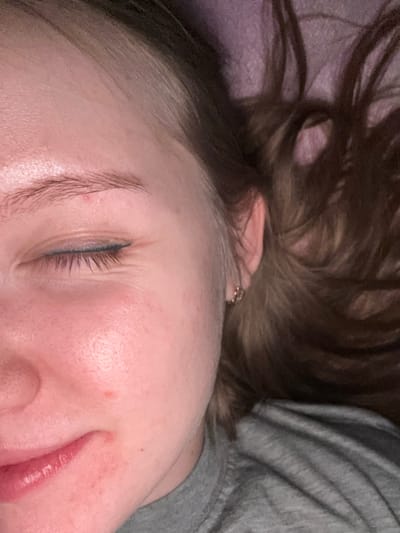 The scene depicts the right side of a female individual's face in a close-up, angled perspective, captured within an indoor environment, understood to be located in Nizhnekamsk, Russia. The subject is positioned lying down, with the right eye closed. A thin line of blue eyeliner is visible along the upper eyelid. Fine lines are present at the outer canthus of the closed eye. The right eyebrow is light brown. The skin appears light-toned with several minor red blemishes on the forehead, right cheek, and near the mouth. The lower portion of the image shows natural pink lips, slightly parted. A small, gold-colored hoop earring is present in the lobe of the right ear. Light brown hair, exhibiting lighter streaks, is fanned out against a textured, dark purple or pink surface, consistent with bedding. A grey fabric garment, identified as a t-shirt, covers the subject's right shoulder and upper chest. Illumination is soft, originating primarily from the left.