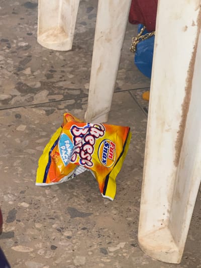 The image shows a scattered bag of Fun Snax Cheese Balls on a terrazzo floor in a domestic setting, possibly within Barnawa, Nigeria. The bag, primarily yellow and orange with blue lettering, lies partially under a weathered, white wooden structure. There is no evidence of people or interaction within the frame. Other unidentified objects are visible in the background.