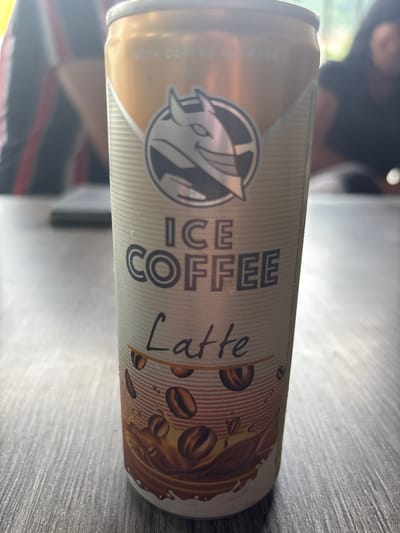 A close-up, slightly angled shot showcases a can of "ICE COFFEE LATTE" with a distinctive logo of a stylized creature. The can is primarily gold and white, with thin horizontal lines creating a textured background on the white sections. The logo, featuring a gray creature with sharp features and possibly horns, is centered on the upper portion of the can. Below the logo, "ICE COFFEE" is printed in large, dark gray block letters, followed by "Latte" in a handwritten-style font. The lower part of the can displays an illustration of coffee beans splashing into a chocolate-colored liquid, with white splashes. Small water droplets are visible on the surface of the can, suggesting it is cold. The can rests on a gray, wooden surface with visible grain. In the blurred background, two individuals are partially visible. One, on the left, wears a red and black striped shirt. The other, on the right, has dark hair and wears a dark t-shirt. The overall setting appears to be indoors, possibly a cafe or restaurant, as indicated by the table surface and the blurred background figures, and the lighting suggests daylight filtering in. The specific location is the city of Karlovo, Bulgaria, though no explicit markers confirm this within the visual frame itself, this information is inferred from external context.
