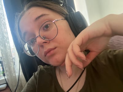 The image displays a young adult female captured indoors in Pushchino, Russia. She is directly facing the camera, wearing black over-ear headphones and round, metal-framed eyeglasses. Her hair, dark in color, is partially visible. A small, reddish mark is present on her forehead. A septal piercing is visible on her nose. She is dressed in an olive-green V-neck shirt, and a silver-toned chain necklace with a small pendant is visible around her neck. Her right hand, exhibiting manicured fingernails with a light, possibly glittered, polish, is positioned with fingers curled against her right jawline and chin. The background on the left side features a brightly lit window covered by white, sheer lace curtains. Partially visible objects on a surface near the window include a white oval item, a plastic bottle, and a black cable. The background on the right is indistinct and light-colored.