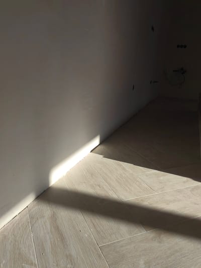 The image shows a corner of an unfinished room with a light-colored wall and light wood-look tile flooring. A diagonal shadow from an unseen object cuts across the floor and part of the wall. The wall has several electrical outlets visible in the background, suggesting it's an interior space. There are no people or specific objects of interest other than the architectural elements and the lighting.