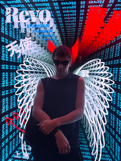 This media file captures a person posing in front of a vibrant, digitally illuminated display in Shanghai. The subject, a person with dark hair and sunglasses, is positioned in the foreground, their body angled towards the viewer. They are wearing a dark, sleeveless top and dark pants. Their right hand is raised, with fingers adorned with rings.
The background is a dynamic and visually rich environment filled with repeating text and abstract shapes. The dominant colors are deep blue, red, and white. Large, stylized white lettering, including "REVO," "SHANGHAI," and what appear to be Chinese characters, are layered and arranged to create a sense of depth and visual interest. Red, abstract forms, resembling flames or sharp geometric shapes, are also prominent. The repeating text "Revo R Revo R" and "BGAO" in a bright blue hue forms a textured backdrop, suggesting a commercial or artistic installation. The overall impression is one of a modern, urban setting, possibly an exhibition or an advertisement, designed to be visually striking. The lighting suggests it could be nighttime or an indoor space with controlled illumination. The composition and the subject's pose suggest a deliberate and artistic presentation, perhaps a promotional image or a personal snapshot capturing a moment at a specific location in Jing'an District, China.