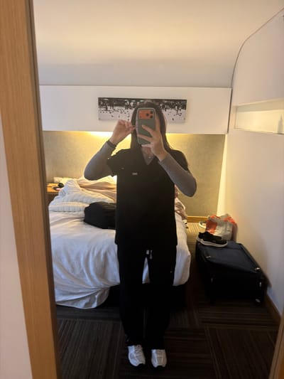 A person stands in a hotel room, reflected in a large mirror. They are wearing dark scrubs, a grey long-sleeved shirt underneath, and white sneakers. The person is holding a smartphone in their right hand and appears to be adjusting something near their ear with their left hand, possibly an earbud. The room has a bed with white linens, a bedside table, and a dark suitcase with a canvas bag on top. The walls are cream-colored, and a piece of art depicting a flock of birds in a winter scene hangs above the bed. The flooring is a patterned carpet in shades of brown and grey. The overall impression is of a tidy but functional hotel room. The image does not provide any specific visual cues to pinpoint the location within Bogota, Colombia.