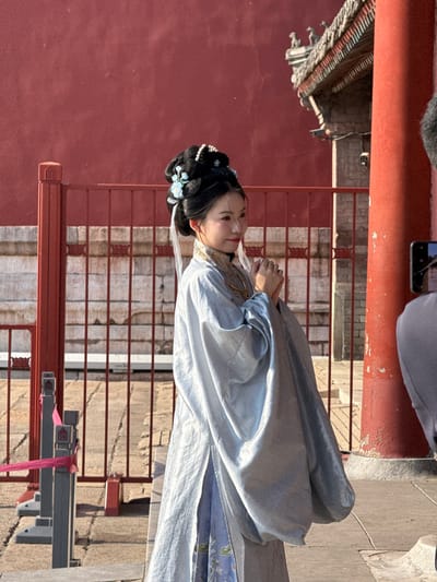 The image shows a woman dressed in traditional Chinese Hanfu clothing standing in front of a red wall with a railing. The location appears to be in the Dongcheng District of China, based on the architectural style. The woman has her hair styled with traditional ornaments. A portion of another person is visible in the upper left corner, and a smartphone is also visible. The railing is made of red-painted metal with a horizontal bar design. The wall is a dark red color, likely part of a traditional building. There is a modern-looking grey fixture with a red ribbon tied around it near the bottom right corner of the image. The ground is paved with stone.