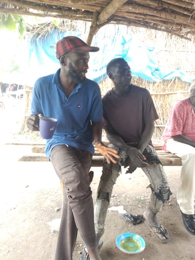 The image shows three people sitting on a wooden bench under a thatched roof structure. The person on the left, wearing a blue polo shirt and a red and black checkered cap, is holding a purple mug. He has a beard and appears to be speaking. To his right, a person in a dark maroon t-shirt and distressed denim jeans is looking to the side, with their hands resting on their lap. The third person, on the far right, is wearing a pink patterned shirt and white pants, also looking to the side. The setting appears to be rustic, with a natural ground surface and what looks like tarpaulin or plastic sheeting behind the people. In the foreground, there is a small blue bowl with some green food and a black plastic bag on the ground. The overall scene suggests a casual gathering in a rural or outdoor location, possibly during daylight hours given the brightness. The clothing and environment are consistent with a warm climate. No discernible text is visible in the image.