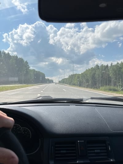 The image shows a view from inside a car driving on a highway. The road is a multi-lane highway with white dashed lines marking the lanes. On either side of the highway, there are dense trees. The sky is blue with large, fluffy white clouds. A portion of the car's dashboard and steering wheel is visible in the foreground, with a hand on the steering wheel. The car's windshield shows some dirt or smudges. The overall environment appears to be a sunny day in a rural or semi-rural area. It is not possible to determine the specific location within Nytva, Russia from the image alone.