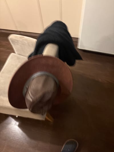 A wide-brimmed, reddish-brown hat with a woven crown is placed upside down on a light-colored, upholstered chair in an indoor setting. A dark, fuzzy fabric, possibly a scarf or shawl, is draped over the back of the chair. The floor is made of dark wood planks. A white vertical surface, likely a wall or appliance, is visible in the background. The lighting appears to be artificial and casting subtle shadows. A dark shoe is partially visible in the lower right corner of the frame. There are no people directly visible in the image. The scene is not specific to any particular location in the city of Orekhovka, Russia.