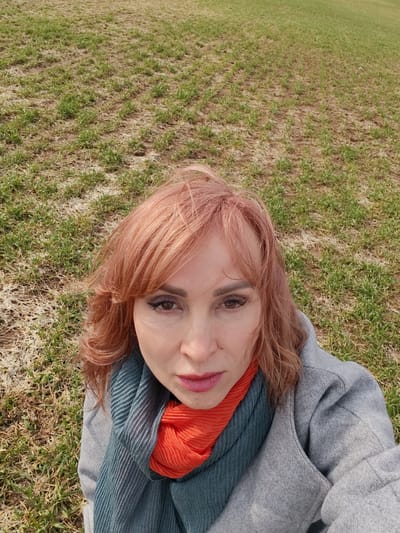 A woman with reddish-blonde hair and light skin stands outdoors. She wears a gray coat with a teal and orange scarf. She looks directly at the camera. The background consists of patchy grass. The exact location within Нечкино, Russia, cannot be determined from the image.
