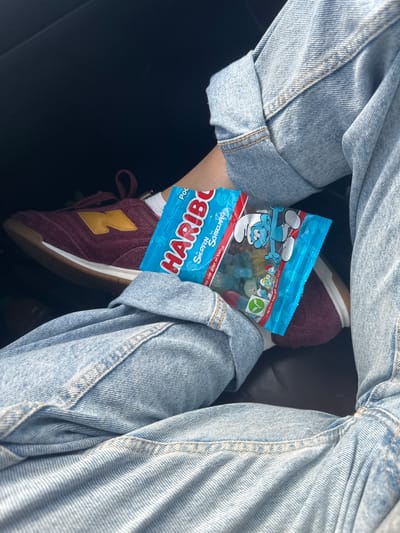 A person's legs are shown in close-up, wearing light blue denim jeans with the cuffs rolled up, and a burgundy New Balance sneaker with a yellow "N" logo. The person is holding a small, bright blue packet of Haribo Smurf gummies. The background is dark and out of focus, suggesting an indoor or shaded environment. There is no visible indication of the location being Wattrelos, France.
