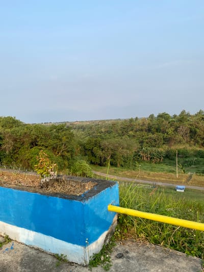 This is a landscape image taken in Bandungan, Indonesia. The photo is oriented vertically. In the foreground, a bright yellow pole rises diagonally from a concrete structure painted with blue and white stripes. The concrete structure appears to be part of a railing or barrier. Beyond this barrier is a densely wooded hillside covered in lush green trees and vegetation. A narrow, paved road runs along the side of the hillside, with a white fence or barrier bordering it in places. In the distance, the land appears flatter and more open, with some patches of dry grass or fields. The sky is a pale, hazy blue, suggesting it may be early morning or late afternoon. The overall scene depicts a natural, somewhat rural environment. There is no visible text in the image.