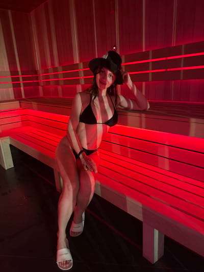 A woman is reclining in what appears to be a sauna, based on the wooden slats and red lighting. She is wearing a black bikini and a hat, as well as white Calvin Klein slides on her feet. The wooden slats are present on the walls and the ceiling. The background is dark with areas of intense red light. The environment is consistent with an indoor sauna setting, possibly in Murino, Russia.
