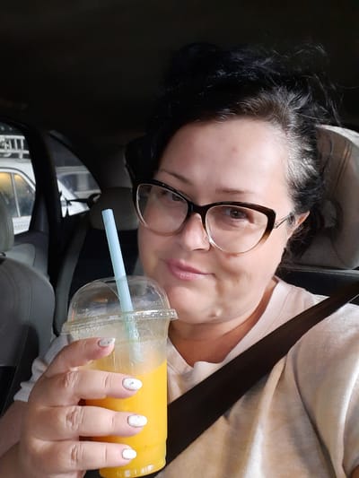 A woman with dark hair and eyeglasses is in a car, wearing a beige-colored shirt and a seatbelt. She is holding a clear plastic cup with a straw, filled with a yellow liquid, likely juice. The environment is the interior of a car. The location cannot be determined based on the image.