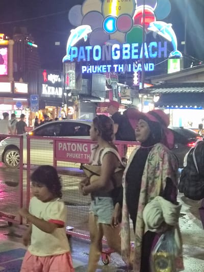 This is a night-time photograph taken in Patong, Thailand. The scene is a bustling street filled with neon signs and people. The image is oriented vertically, with the top of the frame appearing on the left.
The main focus in the foreground and midground is a large, blue neon sign that spells out "PATONG BEACH". A stylized blue dolphin jumps above the sign. To the right of this, a red sign reads "PATONG PHUKET THAILAND". Further down the street, other neon signs are visible, including one that says "KEBAB". There are several people in the scene, some walking and others appearing to be vendors or business owners. The individuals are dressed in casual clothing, and their faces are difficult to discern due to the low light and the orientation of the image.
The setting appears to be a vibrant tourist area, likely a street market or entertainment district. The presence of numerous signs in English suggests it caters to international visitors. The lighting is dominated by the bright neon of the signs, creating a colorful and energetic atmosphere typical of a lively night scene. The ground appears wet, suggesting recent rain or a damp environment. The overall mood is one of activity and commercial vibrancy.