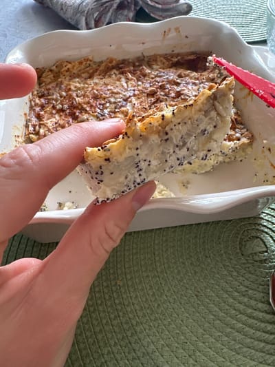 A photograph shows a piece of poppy seed cake or pastry being held up by a person's fingers, having been extracted from a white baking dish that contains the remainder of the cake. The cake has a light-colored topping with a heavy sprinkling of poppy seeds, while the base appears to be a baked, shredded mixture with a golden-brown color. A red utensil is partially visible in the baking dish. The background includes a green woven placemat and the edge of a decorative object with a silver and gray pattern. The location is not evident from the image, therefore, the location details cannot be included.