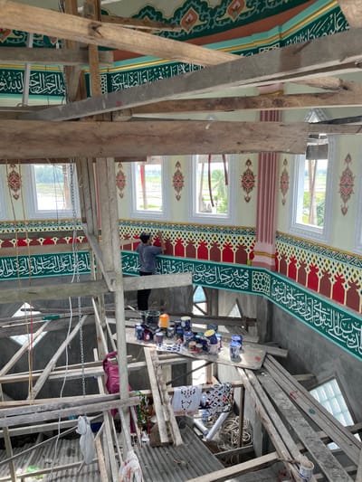 An interior view depicts a male worker painting decorative Islamic patterns within a mosque under construction or renovation in Mns Asan, Indonesia. The individual, wearing a grey t-shirt and dark pants, stands on a wooden scaffolding platform, applying paint to an ornate wall. The walls are painted in cream with intricate green, gold, and pink motifs near the ceiling, and a lower band featuring white Arabic calligraphy on a green background, bordered by red and yellow geometric patterns. Multiple rectangular windows along the upper wall offer a view of a rural landscape with rice paddies and palm trees. Extensive, multi-level scaffolding constructed from rough-cut wooden beams and planks fills the foreground and midground. On a lower platform, numerous paint cans, buckets, brushes, and other painting supplies are visible. The lower levels of the structure appear unfinished, composed of concrete and corrugated metal, with discarded materials, ropes, and a metal chain present.