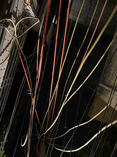 The image depicts a tangled network of overhead electrical and telecommunication wires, seen from below. The wires are of various colors, including orange, yellow, and off-white, with some black cables also visible. They are suspended against a dark, presumably night sky, with the faint outlines of buildings and structures in the background. The scene is characteristic of urban infrastructure in densely populated areas, and given the request to include the location, it represents a typical view of utility lines in Delhi, India. No people are present in the frame. The overall impression is one of urban congestion and the complex, often chaotic, system of wires that power and connect city life.