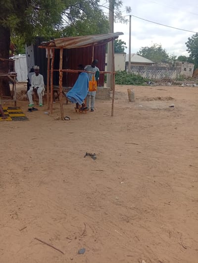 This image captures an outdoor scene in Birnin Kudu, Nigeria, featuring a makeshift barber setup and several individuals on a dirt ground under an overcast sky.

In the center-left, a rudimentary shelter stands, constructed from wooden poles supporting a rusted, corrugated metal roof. Beneath this shelter, a barber, dressed in a light-colored top and trousers, is actively cutting the hair of a client. The client is seated on a low wooden stool and covered with a bright blue cape. The barber holds an orange bag, labeled "Mama's Delight," in his left hand and a green cloth or tool in his right.

To the immediate left of the barber setup, a man wearing a white traditional outfit and a white cap, with bright green sandals, is seated on a low structure, likely a concrete block. His gaze is directed towards the left. Partially visible behind him is another individual in dark attire. Further to the far left, a person's lower arm and hand are visible, resting on a black and yellow patterned mat laid on the ground. A pair of black slippers lies on the dirt near the seated man in white.

The ground consists of reddish-brown, sandy dirt, showing various footprints and scattered small debris including twigs, pebbles, and plastic litter. In the mid-ground to the right, a large, light-colored sack stands upright, and an area of disturbed earth is visible. Piles of green foliage and debris are accumulated in front of concrete block structures and a light-colored building in the background. A dark, corrugated metal building, possibly a container, is situated directly behind the barber's shelter. Further in the background, residential-style buildings with sloped roofs are visible, along with a utility pole and overhead wires. Numerous trees with green canopies frame the scene, particularly on the left and in the distant background. The sky is uniformly grey and overcast.