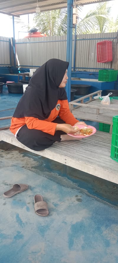 A woman wearing a black hijab and an orange long-sleeved shirt is sitting on a wooden bench and eating from a pink plate. She is in an outdoor covered area with a corrugated metal roof and blue-painted concrete floor. In the background, there are several large black tubs and green crates. Two pairs of sandals are on the floor in front of the bench. The scene appears to be in Puloblang, Indonesia, possibly a fish farm or aquaculture facility, given the presence of tubs and crates commonly used in such environments.