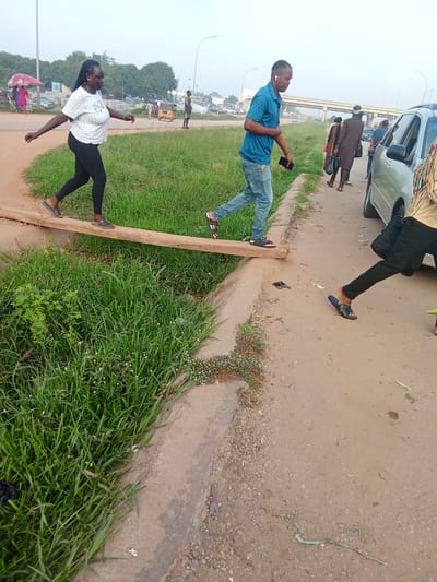 In Kagini, Nigeria, two individuals are depicted navigating a narrow wooden plank stretched across a ditch. On the left, a woman with braided hair, wearing a white checkered t-shirt and black leggings, walks with her arms outstretched for balance. On the right, a man in a blue polo shirt and jeans is in mid-stride, holding a phone and wearing earbuds. The plank is situated on a dirt path next to a grassy area. In the background, a road with cars, an overpass, and some buildings suggests an urban or semi-urban setting. Other pedestrians and vehicles are visible further down the road. The scene captures a moment of everyday life and resourcefulness in crossing an obstacle.