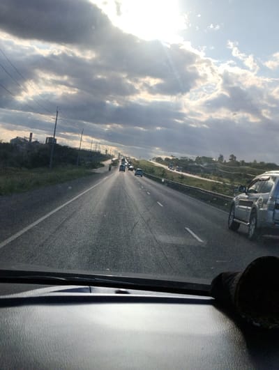 The image shows a view from inside a vehicle traveling on a highway in Athi River, Kenya, at coordinates -1.497 latitude and 37.074 longitude. The highway has a few cars traveling in both directions. The sky is bright with sun shining through the clouds. The vegetation is sparse and the landscape is flat.
