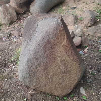 A large, weathered, brownish-grey boulder sits prominently in the foreground, angled towards the right side of the frame. Its surface exhibits a rough texture with visible cracks and a distinct, lighter-colored, curved line running across its upper portion. Several smaller rocks and debris, including dry leaves, twigs, and some small green plants, are scattered around the base of the boulder and in the surrounding dirt ground. In the background, more large rocks are visible, some appearing smoother and more rounded than the central boulder. The overall environment suggests an outdoor setting, possibly a natural area or a rocky landscape within Ribina East, Nigeria. No people or animals are present in the image.