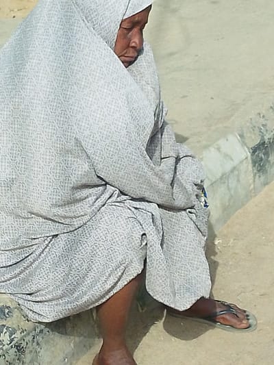 The image portrays a lone individual, an adult female with dark skin, seated outdoors in Babban Giji, Nigeria. She is enveloped in a light grey garment and head covering, both featuring a repeating circular geometric pattern. Her eyes are closed or cast downward, indicating a state of rest or slumber. The garment covers her body entirely except for her lower legs and feet. Her left foot is bare. Her right foot is clad in a dark-strapped flip-flop with a light-colored sole. She is positioned on a light grey concrete or stone surface, which appears to be a curb or a low wall. Adjacent to this seating surface is a light brown, earthy ground. A distinct concrete curb structure is visible, separating the elevated seating area from the ground level. A small, obscured blue and yellow element is faintly visible near her right elbow, beneath the folds of her garment.