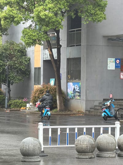 The image depicts an urban street scene in Jiangning District, China. The street is lined with gray buildings featuring barred windows. A tree with green foliage extends across the frame. On the right, a white metal barrier with blue accents is visible. Two individuals are present: one riding a blue scooter near the barrier, and another similarly dressed person on a scooter is located near the building, engaged in some activity by the side of the road. The road surface appears wet. A street sign is mounted on a tree in the middle ground. Posters or signs are affixed to the building. The overall scene suggests a typical urban setting in the district.