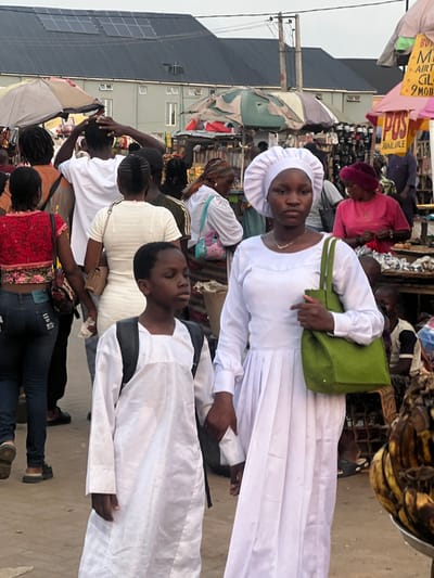 A group of people are walking through an outdoor market. Two people dressed in white robes and hats are prominent in the foreground. The woman has a green purse on her shoulder. The market is bustling with people and vendors. Merchandise and signs are visible among the crowd. The scene appears to be in Nigeria based on clothing and building architecture.