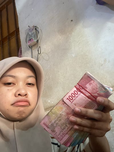 The image shows a close-up of a young woman and a wad of Indonesian Rupiah banknotes. The woman is wearing a light-colored hoodie and is looking up towards the camera. Her face is the primary subject in the lower half of the frame, with her lips slightly parted. In the upper half of the frame, a hand is holding a stack of 100,000 Rupiah banknotes, secured with a red rubber band. The banknotes are pink and feature the text "SERATUS RIBU RUPIAH" and the number "100000" prominently. There are also serial numbers visible on some of the bills, such as "ARF729959".

The background appears to be an indoor setting with a concrete or textured wall. A small object with wires attached, possibly a light fixture or some electronic device, is visible on the wall in the upper left quadrant. The lighting suggests it could be daytime, but the setting is not clearly defined. The overall impression is a casual, personal snapshot. There are no strong indicators of the specific time of day or weather. The emotion conveyed by the woman's expression is neutral or slightly curious. The location context is identified as Ta', Indonesia, which is consistent with the currency shown.